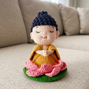 Könnte beinhalten: Eine gehäkelte Figur einer sitzenden Person mit einer dunkelblauen, strukturierten Frisur, einem hell pfirsichfarbenen Gesicht mit geschlossenen Augen und rotem Mund, bekleidet mit einer senfgelben Robe, auf einer rosa Lotusblüte mit grünem Sockel.