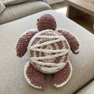 Könnte beinhalten: Ein gehäkeltes Meeresschildkröten-Plüschtier in Altrosa und Weiß. Die Schildkröte hat einen abgerundeten Panzer mit weißen Garnverzierungen, vier Flossen mit weißen Rändern und einen kleinen Kopf mit einem seitlich sichtbaren schwarzen Auge.