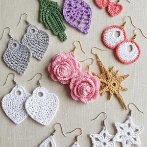 Könnte beinhalten: Eine Sammlung handgefertigter Häkelohrringe in verschiedenen Formen und Farben. Die Ohrringe umfassen Blatt-, Herz- und Blumenmotive in Grün-, Lila-, Rosa-, Weiß- und Grautönen. Jedes Paar hat einen goldfarbenen Haken.