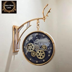 Puede incluir: Un reloj de pared de doble cara con un marco dorado y un borde blanco. La esfera del reloj presenta un diseño de mapa del mundo con engranajes visibles. El reloj está montado en un soporte dorado con un diseño decorativo de asta. El texto "Gem Glow Lane" es visible.