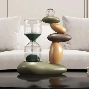 May include: A modern hourglass with a green sand timer, clear glass, and a decorative base. The hourglass is balanced with a stack of smooth, oval-shaped stones in shades of green, brown, and beige. A clear glass sphere sits atop the stone stack.