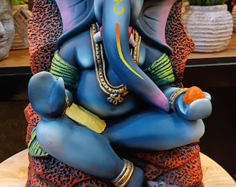 Puede incluir: Estatua azul de la deidad hindú Ganesha, sentado sobre una base con estampado rojo y negro. La figura con cabeza de elefante tiene una trompa amarilla, joyas doradas y sostiene una fruta naranja. Un pequeño ratón gris está en la base.