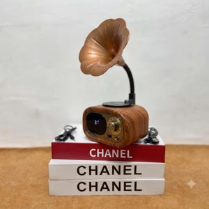 Peut inclure: Une enceinte Bluetooth à effet bois avec un pavillon de gramophone cuivré, un écran numérique affichant BT et un bouton de commande doré. L'enceinte repose sur trois livres empilés blancs et rouges, chacun marqué du texte CHANEL en noir.