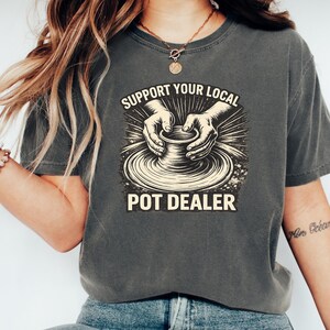 May include: Dark grey t-shirt featuring the text "SUPPORT YOUR LOCAL POT DEALER" in a vintage design. The graphic shows hands shaping a pot on a pottery wheel. The shirt is a crew neck and has a faded look.