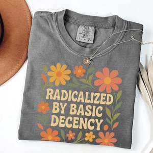 Może przedstawiać: Szara koszulka Comfort Colors z napisem "RADICALIZED BY BASIC DECENCY" w stylu retro, otoczona pomarańczowymi i żółtymi kwiatami oraz zielonymi liśćmi. Srebrny naszyjnik z wisiorkiem leży na koszulce.