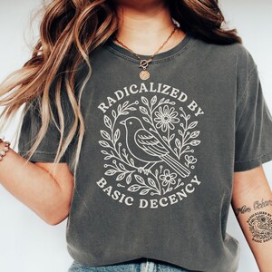 May include: Dark grey t-shirt with the text "RADICALIZED BY BASIC DECENCY" in a white, circular design featuring a bird and floral elements. The shirt is a short-sleeved, crew neck style.