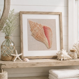 May include: A square framed needlepoint art piece featuring a detailed conch shell in shades of tan, beige, and soft pink, set against a neutral fabric background. The artwork is encased in a light-toned wooden frame with a white mat border.