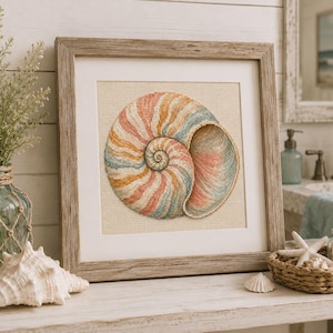 Puede incluir: Una obra de arte en punto de cruz con marco cuadrado que muestra una concha de nautilo con rayas en tonos pastel rosa, azul y amarillo. El bordado está sobre un fondo de tela color crema y dentro de un marco de madera clara.