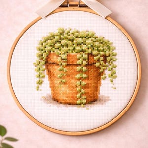 May include: A cross-stitch embroidery piece featuring a potted trailing plant with light green leaves in a terracotta-colored pot, displayed in a circular wooden hoop frame topped with a decorative cream-colored satin ribbon bow for hanging.
