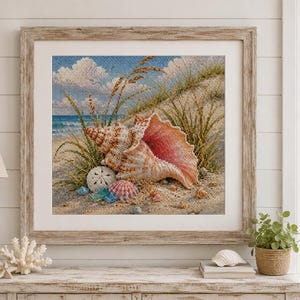 May include: A framed cross-stitch artwork depicting a coastal scene with a large pink conch shell, a white sand dollar, and sea glass resting on a sandy beach. The background features tall dune grass, a blue ocean, and a sky with white clouds.