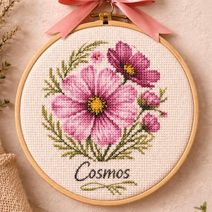 Op de afbeelding: Kruissteekborduurwerk van roze cosmosbloemen met groene bladeren, in een houten hoepel. Het woord "Cosmos" is onder de bloemen geborduurd. Bovenop is een roze lint geknoopt om op te hangen.
