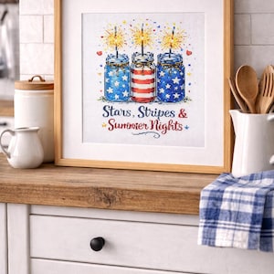 May include: Framed cross-stitch art featuring three mason jars with fireworks. The jars are decorated with stars and stripes in red, white, and blue. The text reads "Stars, Stripes & Summer Nights". The artwork is displayed on a wooden countertop.