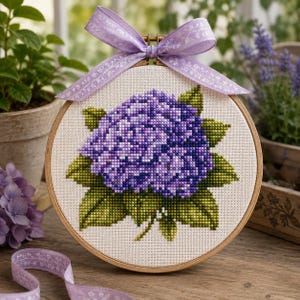 May include: A circular wooden embroidery hoop containing a cross-stitch pattern of a purple hydrangea flower with green leaves on white fabric. A light purple ribbon with a subtle floral pattern is tied in a bow at the top of the hoop.