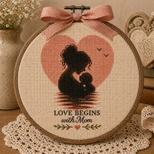 Puede incluir: Un bastidor de bordado de punto de cruz con una silueta de madre e hijo dentro de un corazón rosa. El texto "LOVE BEGINS with Mom" está debajo de la imagen, con un lazo decorativo en la parte superior. El bastidor está enmarcado en madera.