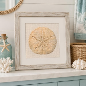 May include: A square framed cross-stitch artwork featuring a tan sand dollar design on a white fabric background. The piece is encased in a weathered, light-colored wooden frame with a white mat, displaying a detailed five-pointed star pattern.