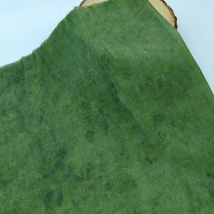 May include: A green fabric with a textured, mottled appearance. The fabric has a slightly worn look, with darker green patches scattered across the surface. The material is folded over a wooden slice, creating a layered effect.