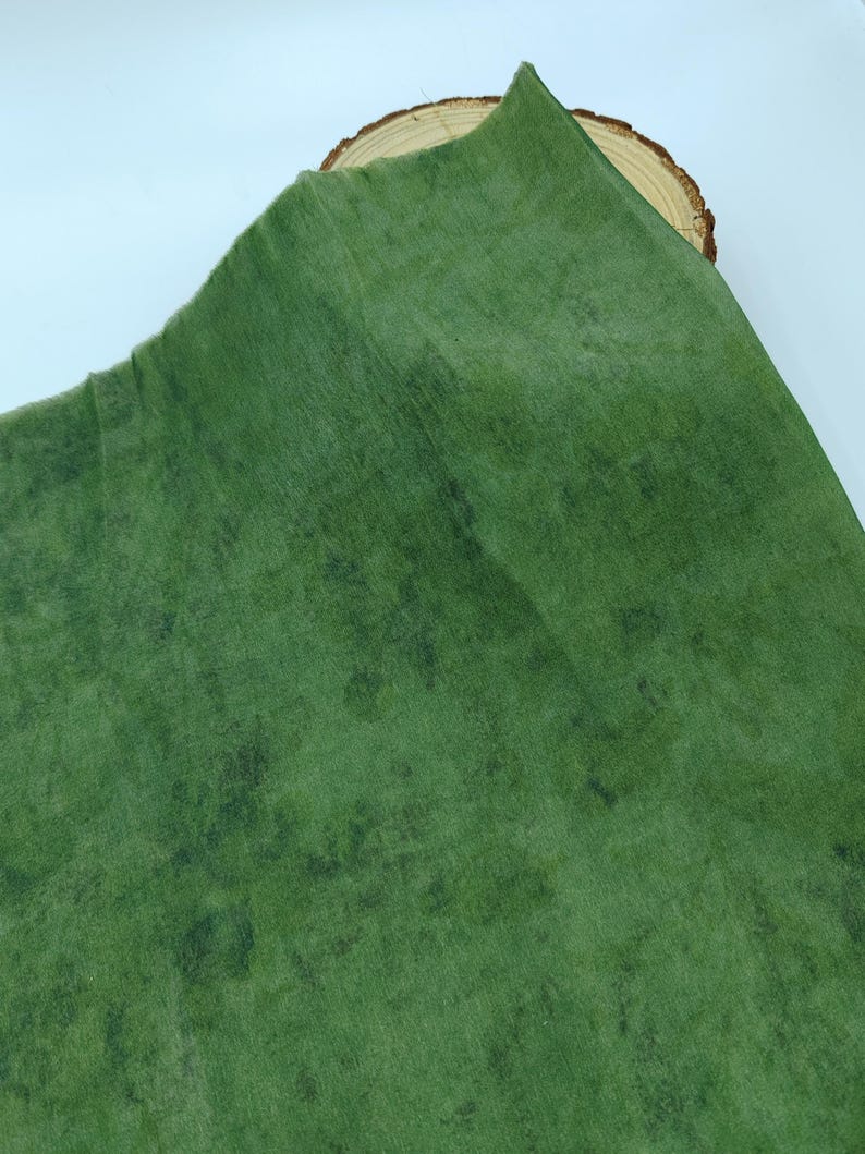May include: A green fabric with a textured, mottled appearance. The fabric has a slightly worn look, with darker green patches scattered across the surface. The material is folded over a wooden slice, creating a layered effect.