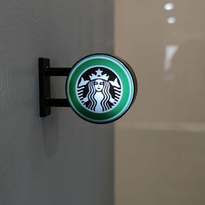 May include: A circular, illuminated Starbucks sign featuring the green border and siren logo. The sign is mounted on a black bracket against a grey wall.