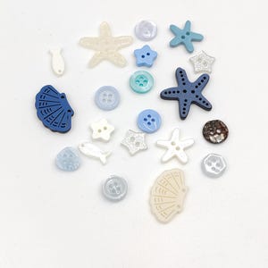Pode incluir: Uma coleção de botões variados em diversas formas e tons de azul, branco e creme. O conjunto inclui estrelas-do-mar, conchas, peixes e botões circulares com dois ou quatro furos, apresentando acabamentos lisos, brilhantes e texturizados.