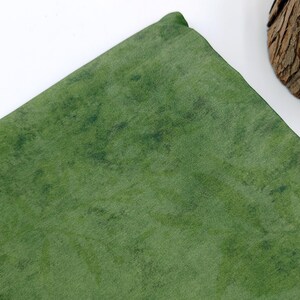 May include: A folded, textured green fabric with darker green mottled patterns. The fabric has a slight sheen, and the corner is rounded. The background is white.