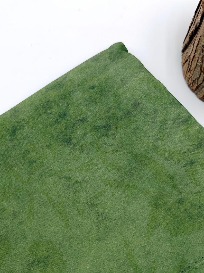 May include: A folded, textured green fabric with darker green mottled patterns. The fabric has a slight sheen, and the corner is rounded. The background is white.