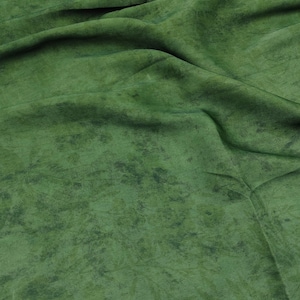 May include: Close-up of a green fabric with a mottled texture. The fabric has a soft, velvety appearance with darker green patches scattered across the surface. The folds and creases in the fabric create shadows and depth.