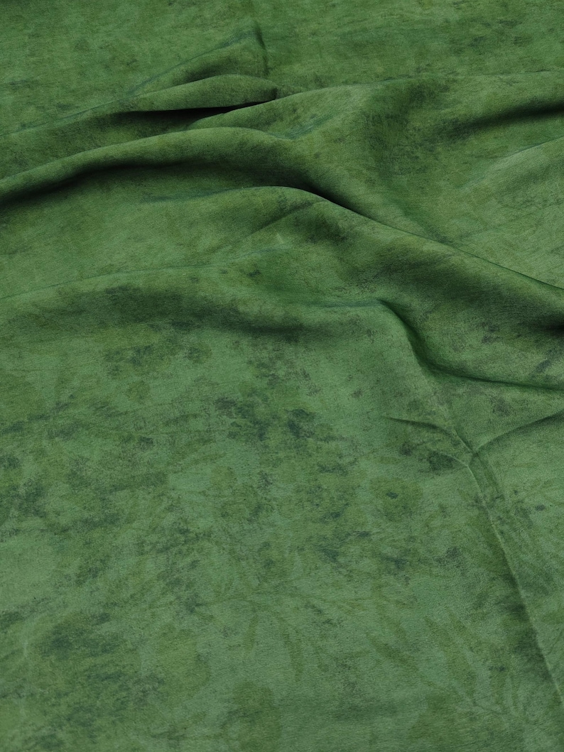 May include: Close-up of a green fabric with a mottled texture. The fabric has a soft, velvety appearance with darker green patches scattered across the surface. The folds and creases in the fabric create shadows and depth.