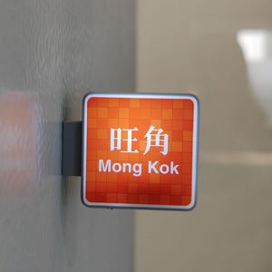 May include: A square sign with the words "旺角" and "Mong Kok" in white text on an orange background. The sign is mounted on a gray metal bracket and attached to a textured gray wall.