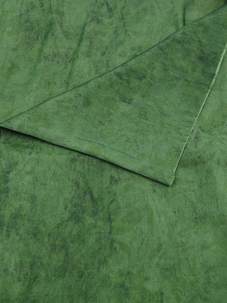 May include: Close-up of green fabric with a marbled effect. The material has a folded corner, showing a lighter green shade. The fabric appears soft and flowing, potentially for clothing or home furnishings. The texture is detailed.