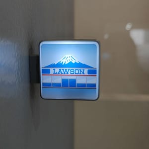 May include: A square, illuminated sign with the Lawson logo, featuring a mountain and the word "LAWSON" in blue. The sign is mounted on a gray surface.
