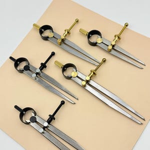 May include: A collection of five metal dividers with silver legs, black adjustment rings, and brass-colored adjustment screws. The dividers are arranged on a light tan surface, showcasing their precision and craftsmanship. These tools are ideal for precise measurements and drawing.