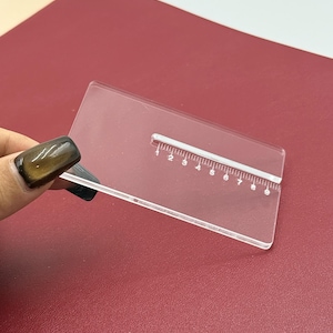 May include: A clear, rectangular acrylic ruler with white markings, measuring up to 20 cm. The ruler is held over a burgundy surface. The ruler is designed for precise measurements.