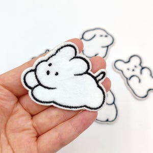 Könnte beinhalten: Weiße, wolkenförmige, gestickte Aufnäher mit schwarzen Umrissen und kleinen schwarzen Punkten. Die Aufnäher haben ein weiches, strukturiertes Aussehen. Mehrere Aufnäher sind sichtbar, einer wird in einer Hand gehalten.