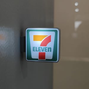 May include: Illuminated square sign for 7-Eleven, featuring a white background with a green border. The logo includes a yellow bar, a red curved shape, and the word "ELEVEN" in teal.