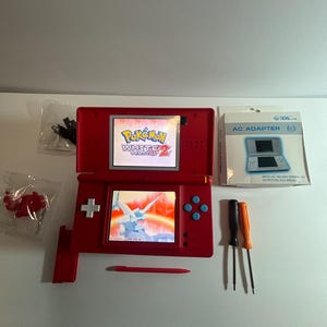 May include: A red Nintendo DS Lite handheld gaming console displaying the Pokemon White Version 2 title screen. Included are a matching red stylus, a white AC adapter box, two small screwdrivers, a red plastic component, and a small black cable.