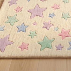 May include: A rectangular area rug with a cream-colored base featuring a repeating pattern of tufted stars in various sizes and pastel shades, including light pink, mint green, lavender, and soft blue, distributed across the entire surface.