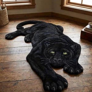 Puede incluir: Una alfombra negra con forma de pantera y ojos verdes, sobre un suelo de madera. La alfombra es de un material suave y texturizado, con detalles en las patas, cola y cara de la pantera.