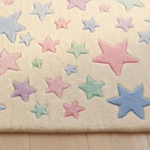May include: A cream-colored rug featuring a repeating pattern of five-pointed stars in various sizes and pastel shades, including light blue, soft pink, mint green, and lavender. The stars have a textured, raised appearance against the flat base.