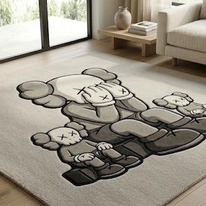A luxurious hand tufted wool area rug displayed in a bright modern living room with hardwood floors. The rug features a large streetwear-style cartoon bear character holding two smaller companion figures, rendered in warm cream, medium grey, and deep charcoal tones. The plush wool pile creates rich texture and depth. Soft natural light highlights the dimensional design. A beige sofa and indoor plant complete the minimalist, hype-culture-inspired interior setting.