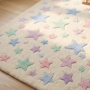 May include: A rectangular area rug with a cream-colored base featuring a repeating pattern of five-pointed stars in pastel shades of pink, blue, purple, and mint green. The rug has a textured, plush surface and is laid out on a wooden floor.
