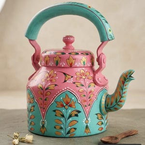 Puede incluir: Una tetera de metal con una base rosa y motivos florales pintados a mano en naranja, amarillo y verde. La parte inferior y el asa están pintadas en azul turquesa brillante. La tapa tiene un pomo rosa y el pico es curvo con patrones florales a juego. El texto Gaurangidecor es visible en la esquina inferior derecha.