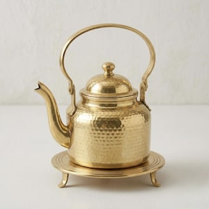 Op de afbeelding: Een theepot van gepolijst messing met een gehamerde textuur, een gebogen tuit en een afgerond deksel met een kleine knop. De theepot heeft een hoog, gebogen handvat en staat op een bijpassende ronde messing standaard met drie korte pootjes.