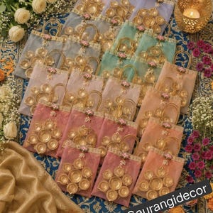 May include: A collection of sheer organza gift bags in pastel colors, each featuring a gold-toned metal diya lamp arrangement on the front. The bags are adorned with a decorative pink lotus flower accent and gold trim, arranged on a patterned blue fabric.