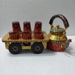 Peut inclure: Un service à thé décoratif avec une théière peinte à la main et un chariot de service avec des verres. La théière est rouge, dorée et verte avec des motifs détaillés. Le chariot contient six verres et a des roues noires.