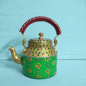 Op de afbeelding: Een decoratieve metalen theepot met een felgroene basis en gele en rode bloemmotieven. Het bovenste gedeelte is goudkleurig met gedetailleerde bloemversieringen en het gebogen handvat is donkerrood geschilderd met witte accenten.