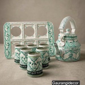 May include: A white tea set featuring a kettle and six cups decorated with intricate green floral patterns. The set includes a rectangular tray with six circular cutouts for the cups. The text Gaurangidecor is visible in the bottom right corner.