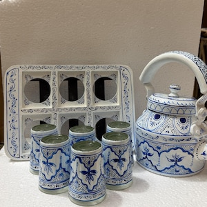 May include: A white ceramic tea set featuring a blue hand-painted pattern. The set includes a kettle with a curved handle and lid, four cylindrical cups, and a rectangular tray with six circular cutouts for holding the cups securely.