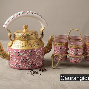 Könnte beinhalten: Eine rosa-goldene Teekanne aus Metall mit weißem Griff und Blumenmustern, dazu vier passende rosa Gläser in einem goldfarbenen Drahtgestell. Die Kanne und die Gläser sind mit handgemalten Verzierungen versehen. Text: Gaurangidecor.
