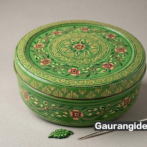 Puede incluir: Un recipiente redondo de metal verde con motivos florales pintados a mano en tonos rosa, amarillo y blanco. La tapa y los laterales están decorados con enredaderas y hojas detalladas. Un pequeño dije en forma de hoja y un pincel fino cerca.