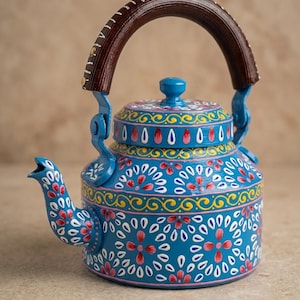 Op de afbeelding: Een felblauwe metalen theepot met ingewikkelde, handgeschilderde bloem- en geometrische patronen in roze, witte en gele tinten. De theepot heeft een bijpassend deksel met een kleine knop en een gebogen, donkerbruin handvat met textuur.